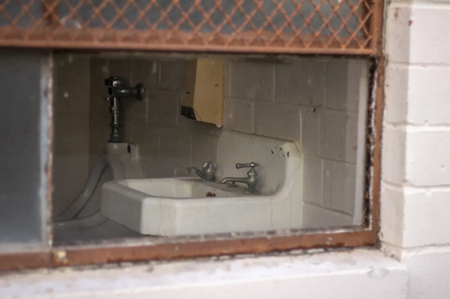 A white porcelain sink and exposed plumbing seen through a broken concrete-block wall opening into a derelict bathroom