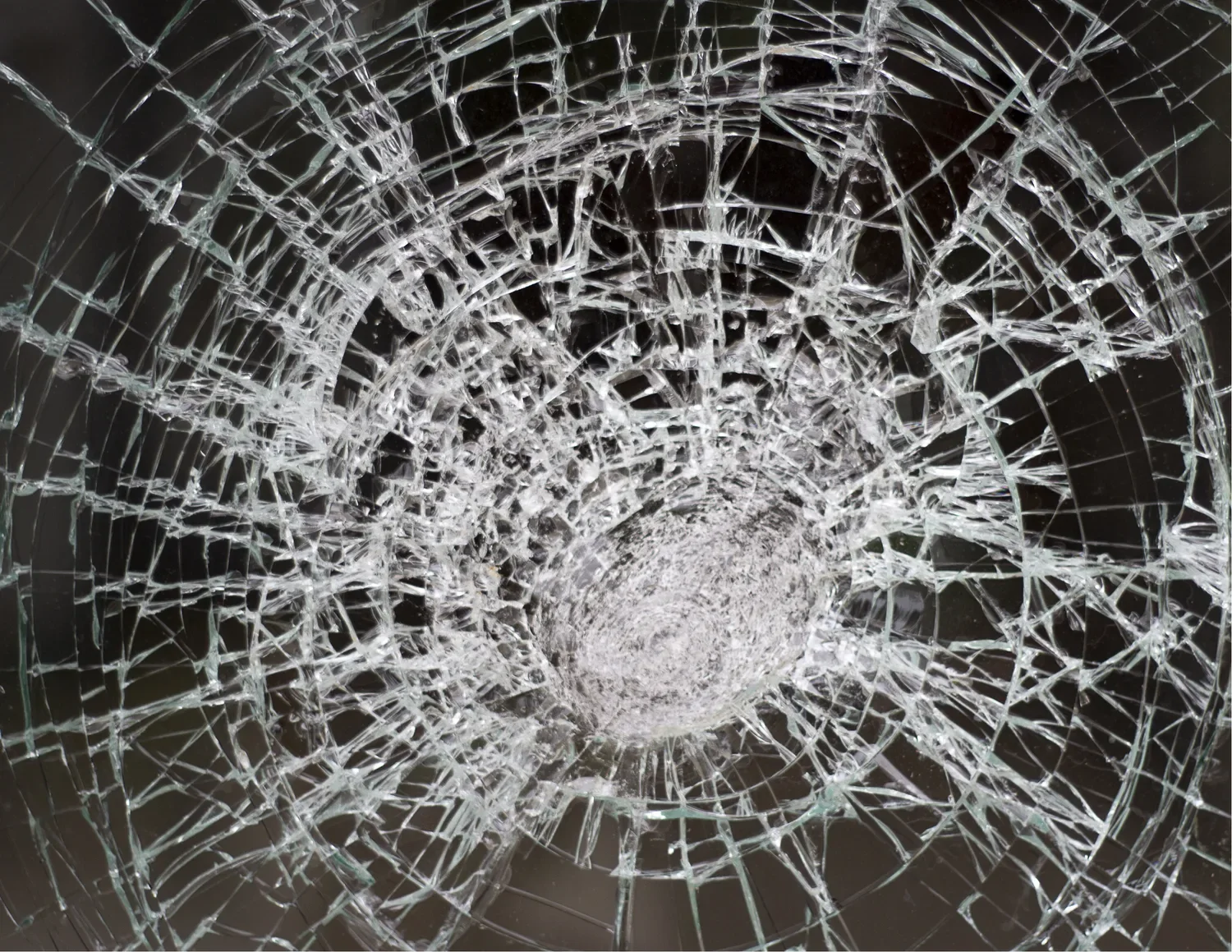 A close view of shattered safety glass, the spiderweb of cracks radiating from a single impact point against a dark interior