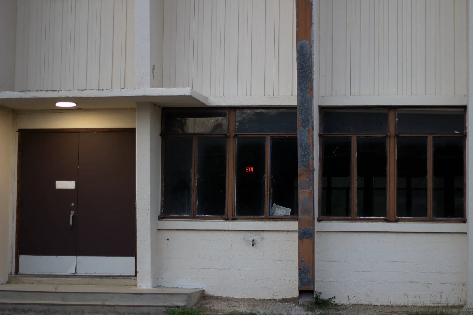 A dark brown metal door beside a band of blacked-out windows in a white painted school wall, weeds at the threshold