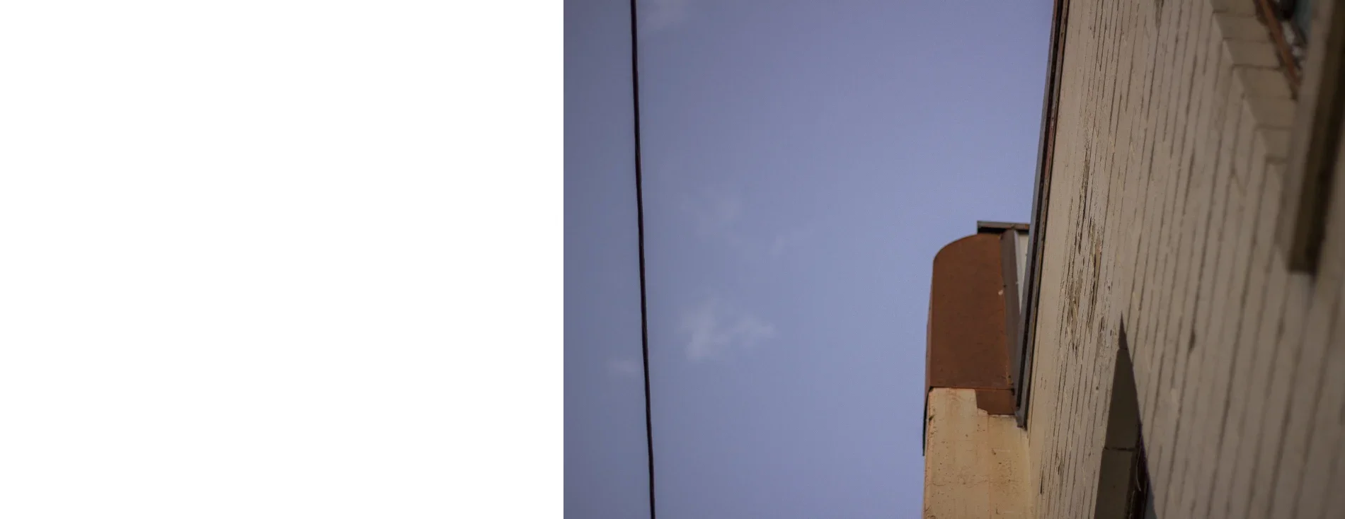 A spread from Lindsey's Crown — the verso left empty, a photograph filling the recto and crossing back over the gutter: a steep low angle up the school's weathered clapboard wall and rusted parapet against a dusk-blue sky, a single service wire cutting the frame