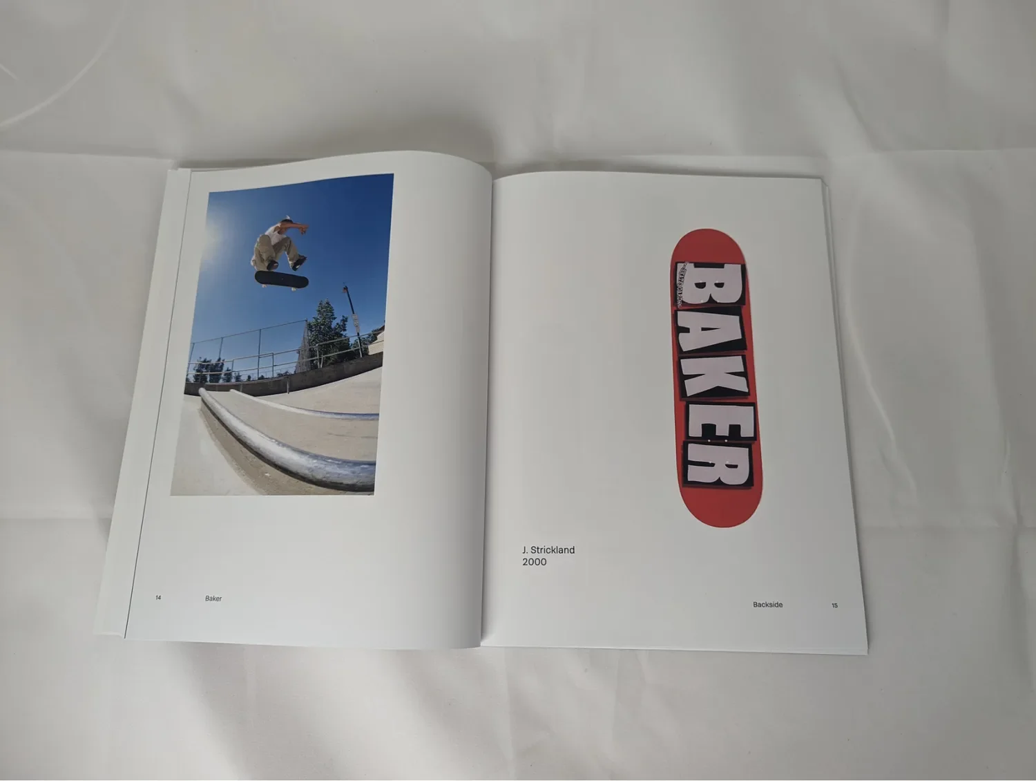 The printed catalog open on a white sheet to pages 14 and 15 — a skater mid-air over a rail on the left, a red Baker deck on the right captioned J. Strickland 2000 — the perfect-bound spine lying almost flat
