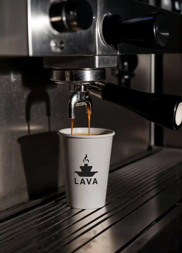 LAVA branded paper coffee cup in the foreground with an espresso machine pulling a shot behind it, the black icon and wordmark on a white cup
