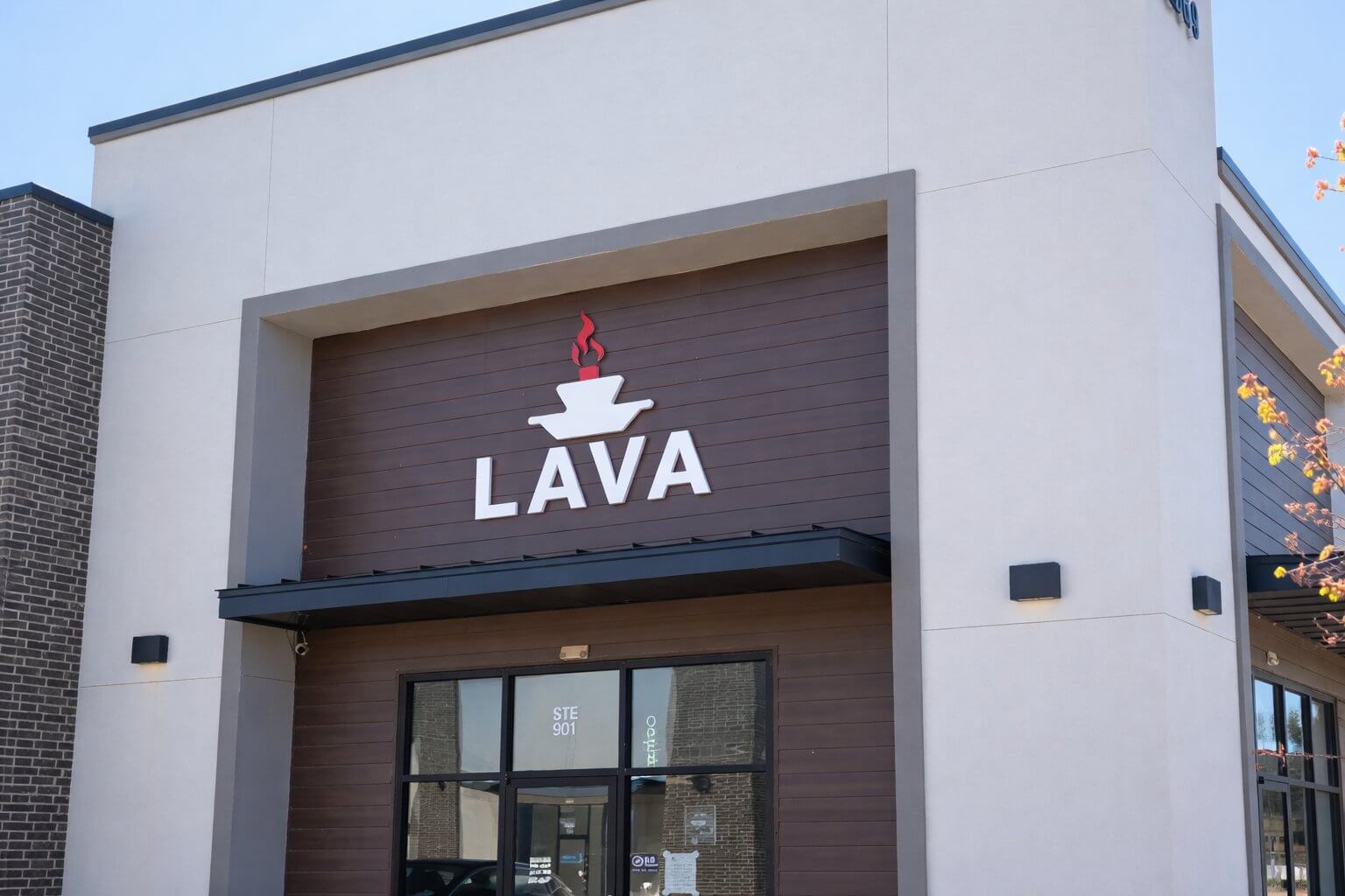 LAVA storefront entrance in daylight, white dimensional letters and color mark mounted above the glass door on a dark wood panel facade